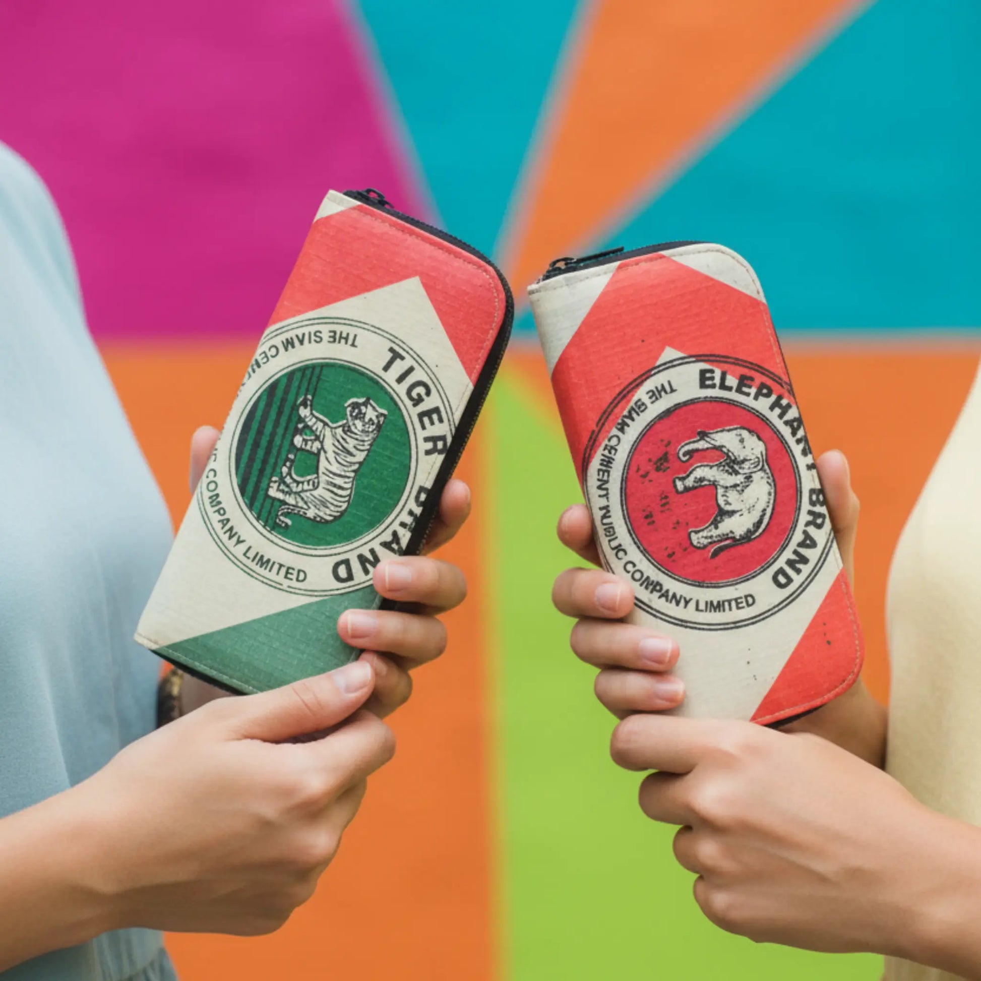 Two hands holding red and green zippered wallets with 'Tiger Brand' logo and Elephant Brand logo against a colorful background from Tuktuk Trading